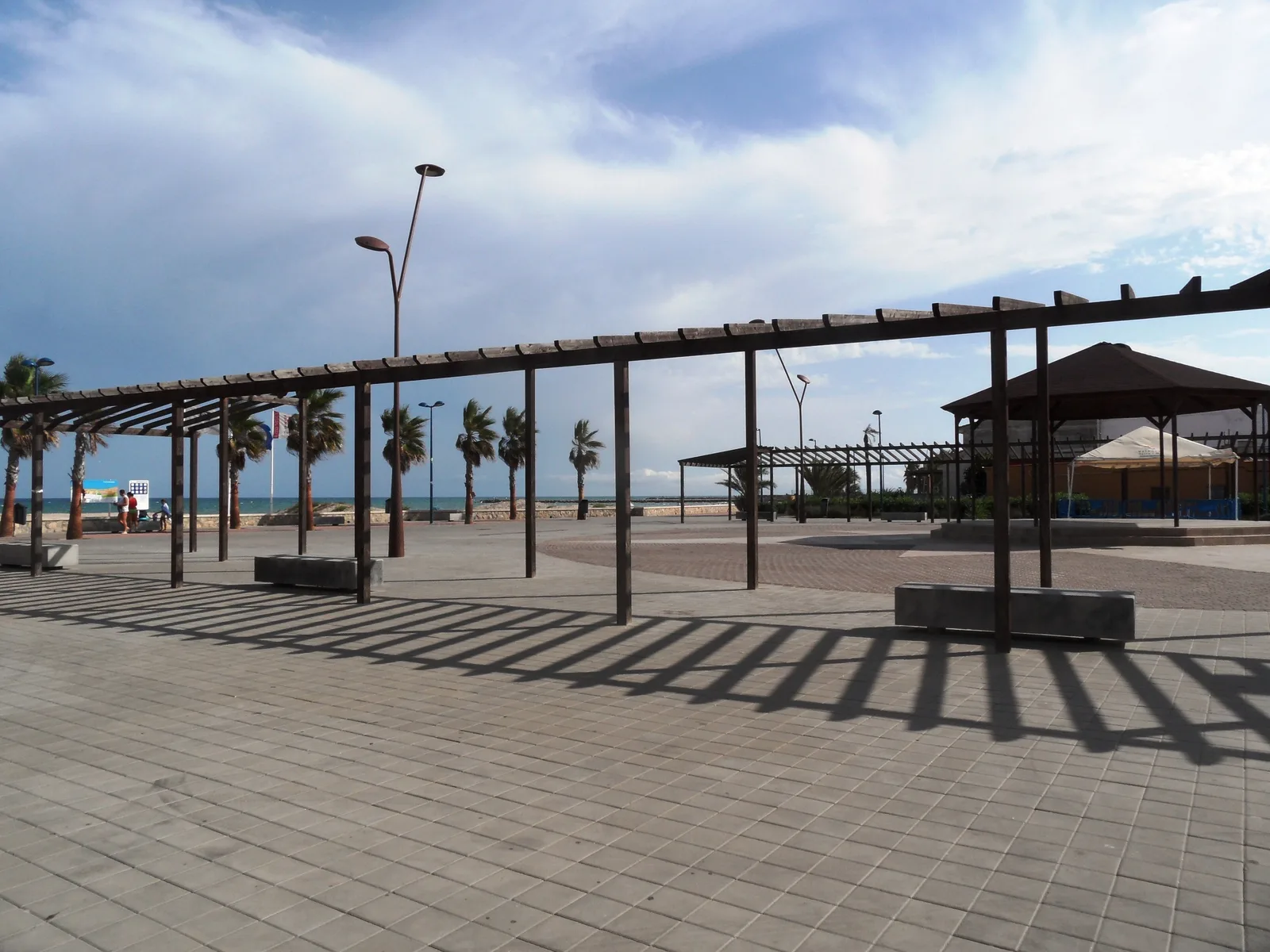 Plaza pavimentada junto al mar con una pérgola de madera en primer término, hileras de palmeras, bancos de piedra, farolas y un pabellón de cubierta piramidal; al fondo se ve el mar y varias personas pequeñas junto a la orilla.